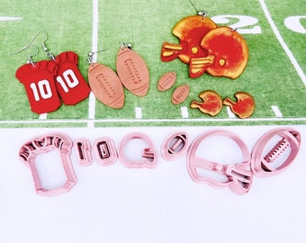 May include: A set of football-themed polymer clay earrings, including a football helmet, a football, and a jersey with the number "10" on it. The earrings are displayed on a green and white surface with pink cookie cutters shaped like footballs and helmets.