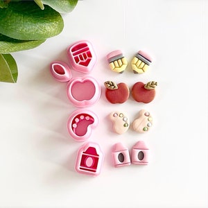 May include: A set of pink plastic cookie cutters in various shapes, including an apple, a pencil, a paint palette, a footprint, and a schoolhouse. The cutters are arranged on a white surface.