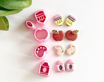 May include: A set of pink plastic cookie cutters in various shapes, including an apple, a pencil, a paint palette, a footprint, and a schoolhouse. The cutters are arranged on a white surface.