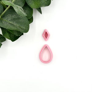 May include: Two pink polymer clay cutters in the shape of a diamond and a teardrop. The teardrop cutter is larger than the diamond cutter. The cutters are on a white background with green leaves in the upper left corner.