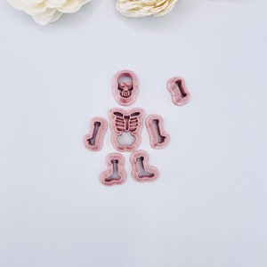 May include: A set of seven pink plastic cookie cutters shaped like a skeleton. The cutters include a skull, ribcage, and four long bones.