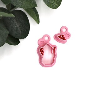 May include: Two pink plastic cookie cutters, one shaped like a teapot with a heart on top, and the other shaped like a cupcake with a heart on top.