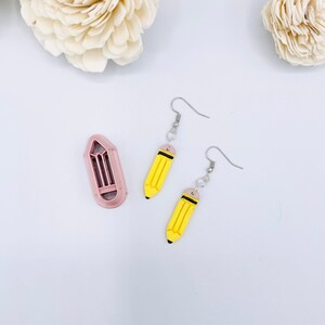 May include: A pair of yellow pencil earrings with black lines and pink eraser details. The earrings are hanging from silver hooks. A pink pencil-shaped cookie cutter is also shown in the image.