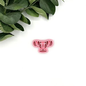 May include: Pink plastic cookie cutter shaped like a moose with large antlers. The moose has a simple cartoon face with eyes, a nose, and a mouth.