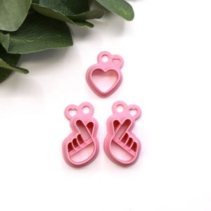 May include: Three pink plastic cookie cutters: two are shaped like hands making the peace sign, and one is shaped like a heart with a small heart on top.