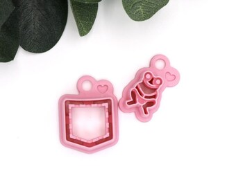 May include: Two pink plastic cookie cutters, one shaped like a frog and the other shaped like a pocket with a heart on top.