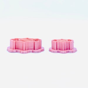 May include: Two pink plastic cookie cutters shaped like a flower. The larger cutter is in the background and the smaller cutter is in the foreground.