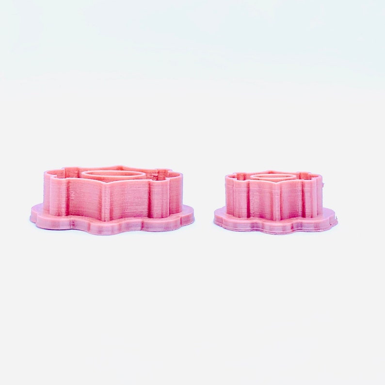 May include: Two pink plastic cookie cutters shaped like a flower. The larger cutter is in the background and the smaller cutter is in the foreground.