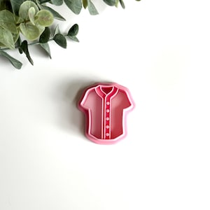May include: Pink plastic cookie cutter shaped like a baseball jersey with buttons.