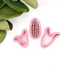 May include: Three pink plastic cookie cutters shaped like corn on the cob. The cutters are on a white background.