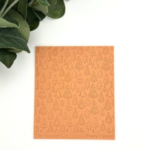 May include: A close-up of a brown silicone baking mat with a repeating pattern of pine trees. The mat has the text "The Sea Salt Co" and "Piney Forest" printed on it.