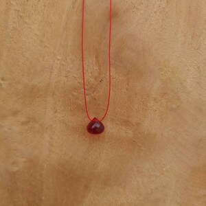 Puede incluir: Un colgante de piedra roja en un collar de cuerda roja. El colgante tiene forma de lágrima y está colgando sobre un fondo de madera.