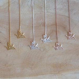 Op de afbeelding: Vijf delicate kettingen met origami-kraanvogel hangers. De kraanvogels zijn gemaakt van zilver, goud en roségoud. De kettingen hangen aan een houten oppervlak.