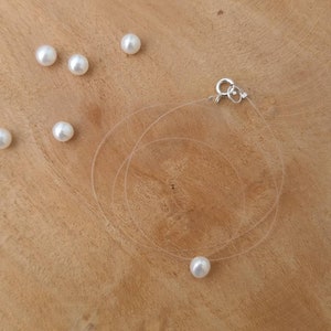Può includere: Una collana di perle singola su una corda trasparente con una chiusura argentata. La collana è appoggiata su una superficie di legno con cinque perle sciolte sparse intorno.