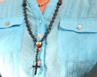 May include: A black beaded necklace with a heart pendant and a cross charm. The necklace is worn by a person wearing a light blue shirt.