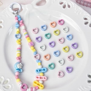 Peut inclure: Un porte-clés à perles avec une variété de perles aux couleurs pastel, dont des perles en forme de cœur et carrées. Un petit personnage de dessin animé est attaché au porte-clés. Des perles en forme de cœur de différentes couleurs sont éparpillées.
