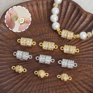 Könnte beinhalten: Eine Auswahl an magnetischen Schmuckverschlüssen in Gold- und Silbertönen, besetzt mit klaren Strasssteinen. Die Verschlüsse werden auf einer Holzoberfläche gezeigt, mit einer Nahaufnahme des Magnetanschlusses. Eine Perlenkette ist ebenfalls sichtbar.