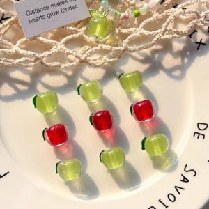 Op de afbeelding: Een verzameling kleine, appelvormige kralen in rood en groen, met een groene appelhanger aan een kralenketting. De kralen zijn gerangschikt op een wit bord met tekst. Een klein kaartje leest "Distance makes the hearts grow fonder."