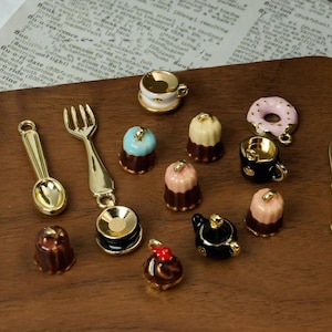 Puede incluir: Una colección de miniaturas doradas de cubiertos y alimentos, que incluyen una cuchara, un tenedor, un cuchillo, tazas de té, rosquillas y pasteles. Los dijes son de varios colores, como rosa, azul y marrón, y están dispuestos sobre una superficie de madera.