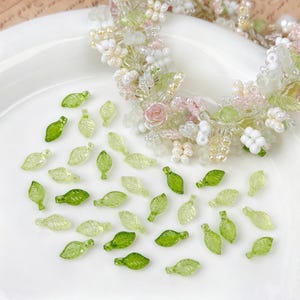 Könnte beinhalten: Ein zarter Blumenkranz mit rosa, weißen und grünen Perlen und Blumen. Kleine, durchscheinende grüne blattförmige Perlen sind um den Kranz herum verstreut. Auf einer weißen Oberfläche.