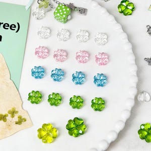 Puede incluir: Una colección de cuentas en forma de trébol de colores claros, rosa, azul y verde. Un dado verde con lunares blancos y un pequeño dije están unidos a una cadena. Las cuentas están dispuestas sobre un plato blanco con volantes.