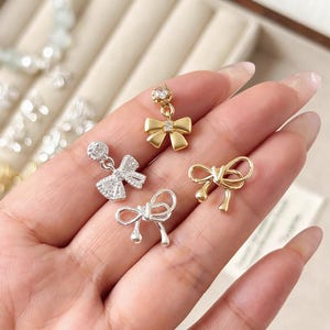 Puede incluir: Una colección de joyas en forma de lazo en tonos dorados y plateados. Un pendiente de lazo dorado presenta un cristal colgante. Otros lazos son de oro macizo o plata, algunos con detalles de cristal. Las joyas se exhiben en una mano.