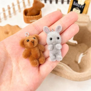 Puede incluir: Un charm de oso de peluche marrón y un charm de conejo gris con ojos negros y un lazo plateado para colgar. El oso de peluche está hecho de un material suave y difuso. El charm de conejo tiene orejas largas.