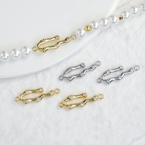 May include: Gold and silver-toned jewelry clasps with a curved design, each adorned with small, clear gemstones. The clasps are displayed near a pearl necklace, showcasing their use in jewelry making and design.