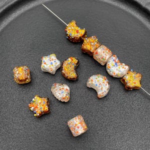 May include: A collection of colorful glass beads in various shapes, including stars, moons, and cubes. The beads are adorned with small, speckled details in shades of orange, blue, and white. Some beads are strung on a thin metal wire.