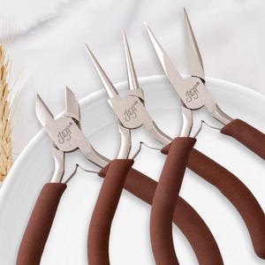 Può includere: Sono presentate tre pinze per gioielli argentate con manici marroni. Le pinze hanno la scritta "Jeff" stampata. Il testo "Imported High-Carbon Steel Jewelry Pliers" è in alto. È presente anche il testo "Double-Quenched Jaw Treatment".