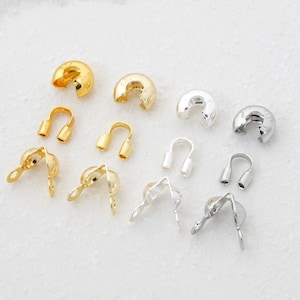 May include: An assortment of jewelry findings in gold and silver tones. The findings include crimp covers, end caps, and cord ends, all designed for jewelry making and crafting. The pieces are arranged on a white surface.