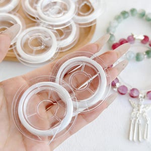 Puede incluir: Bobinas de plástico transparente llenas de hilo blanco para abalorios. Se ven varias bobinas, algunas sujetas con la mano, otras en una bandeja de madera. Una pulsera de cuentas con piedras rosas y verdes está al fondo.
