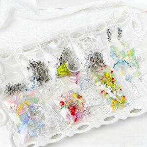 May include: A selection of craft supplies, including beads, pins, and charms, packaged in clear plastic bags. The assortment features various colours and shapes, with some items resembling butterflies and flowers. The items are arranged on a white, decorative tray.