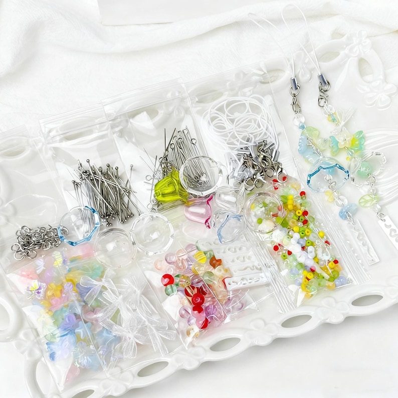 May include: A selection of craft supplies, including beads, pins, and charms, packaged in clear plastic bags. The assortment features various colours and shapes, with some items resembling butterflies and flowers. The items are arranged on a white, decorative tray.