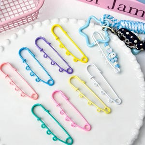 May include: A collection of colorful safety pins in various shades, including pink, blue, yellow, and green. Each pin has a decorative design with small circular accents. A keychain with a star and bunny charm is also present.