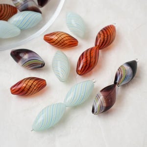 May include: Artisan glass beads in a variety of colours and patterns. Some are translucent light blue with thin blue stripes, others are deep orange with brown stripes. Several are connected in pairs.