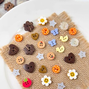 May include: A collection of decorative buttons in various shapes and colors, including flowers, stars, hearts, and circles. The buttons are made of wood, plastic, and shell, and are arranged on a burlap surface.
