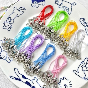 May include: A collection of colorful lanyards with silver-colored clasps. The lanyards are in various colors, including red, green, yellow, white, blue, purple, and pink. The lanyards are arranged on a white plate with a dog pattern.