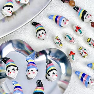 May include: A collection of colorful ceramic beads with whimsical designs. The beads feature a clown-like face with a striped hat in various colors, including black, yellow, blue, and pink. Some beads are oval-shaped with floral or character designs.
