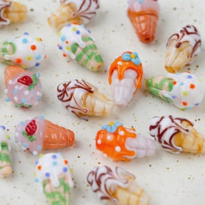 May include: A collection of colorful glass ice cream cone beads. The beads feature various designs, including sprinkles, drizzles, and fruit toppings. The cones are in shades of orange, brown, and white, with the ice cream in pink, white, and green.