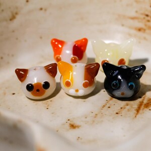 Könnte beinhalten: Fünf katzenförmige Glasperlen in verschiedenen Farben. Die Perlen haben Weiß, Orange, Gelb, Braun und Schwarz. Jede Perle hat einen einzigartigen Gesichtsausdruck und ein einzigartiges Design mit sichtbaren Augen und Ohren. Die Perlen sind auf einer hellen Oberfläche angeordnet.