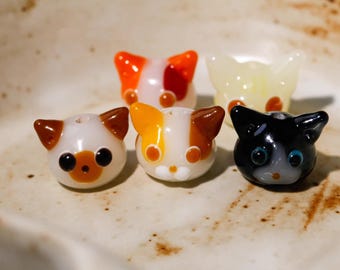 May include: Five colorful cat-shaped glass beads. The beads feature a variety of colors, including white, orange, yellow, brown, and black. Each bead has a unique facial expression and design, with visible eyes and ears. The beads are arranged on a light-colored surface.