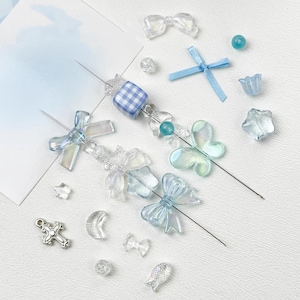 May include: A collection of craft beads and charms in shades of blue and clear. The selection includes bows, butterflies, and a cross pendant. Various shapes are present, including spheres, stars, and flowers. Two beaded skewers are also visible.