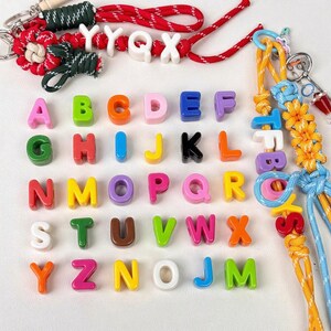 Peut inclure: Un assortiment de perles alphabet colorées dans diverses nuances, dont le rose, le vert et le bleu. Les perles sont disposées en rangées, certaines étant enfilées sur des cordons. Des porte-clés avec des nœuds et des perles décoratifs sont également visibles.