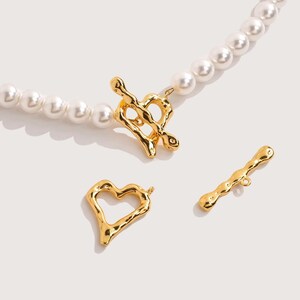 Puede incluir: Un collar de perlas con un cierre dorado en forma de corazón y dos componentes dorados adicionales en forma de corazón y barra. El collar presenta perlas blancas y redondas. Los elementos dorados tienen un diseño texturizado y orgánico.