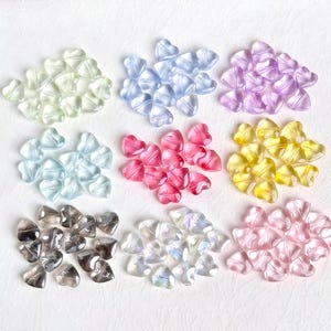 May include: Nine piles of heart-shaped beads in various colours, including light green, blue, purple, light blue, pink, yellow, grey, iridescent, and pink. The beads are translucent and appear to be made of plastic or glass.