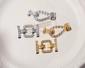 May include: Close-up of four jewelry clasps in silver and gold tones. The clasps are encrusted with clear, sparkling gemstones. Two clasps are silver, and two are gold. The clasps are designed with a hook and eye closure.