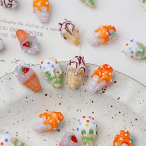 Può includere: Una collezione di ciondoli a forma di cono gelato in miniatura e colorati. I ciondoli presentano vari disegni, tra cui fragola, cioccolato e zuccherini. I coni sono nelle tonalità dell'arancione, del rosa, del bianco e del verde, con colature marroni e bianche. Ideale per la creazione di gioielli.
