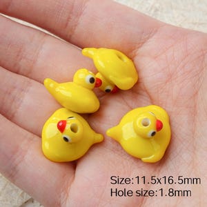 May include: A selection of yellow duck-shaped beads with black and red details. These glossy beads have a hole for threading. The image text indicates the size is 11.5x16.5mm and the hole size is 1.8mm.