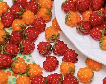 May include: A collection of red and orange glass beads shaped like raspberries. The beads have green stems and are scattered on a surface, with some in a white dish. The beads are translucent and appear to be made of glass.